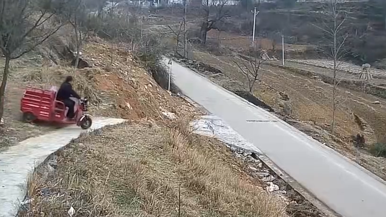 A still frame showing the motorized tricycle traveling along the narrow concrete path with the rural hillside and road visible.