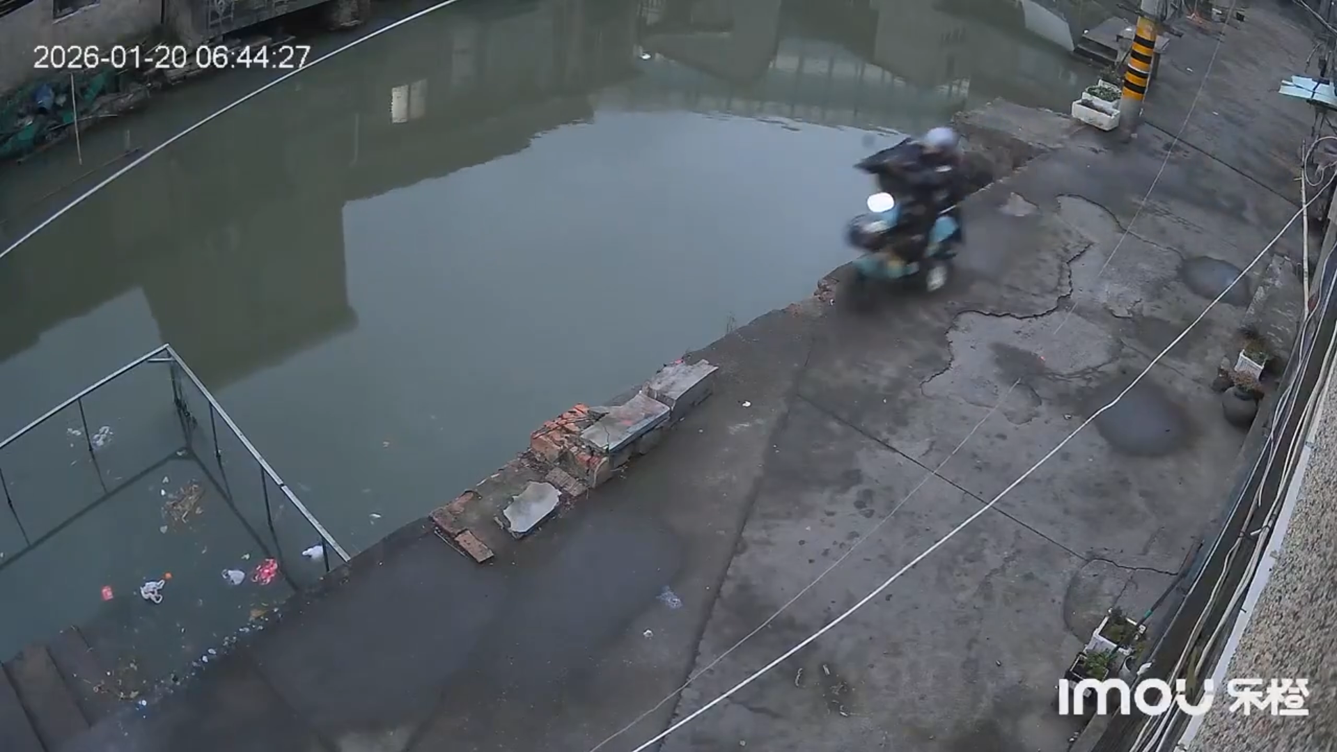 A wide CCTV frame showing the narrow street and canal moments before the scooter enters the water, without close-up imagery.