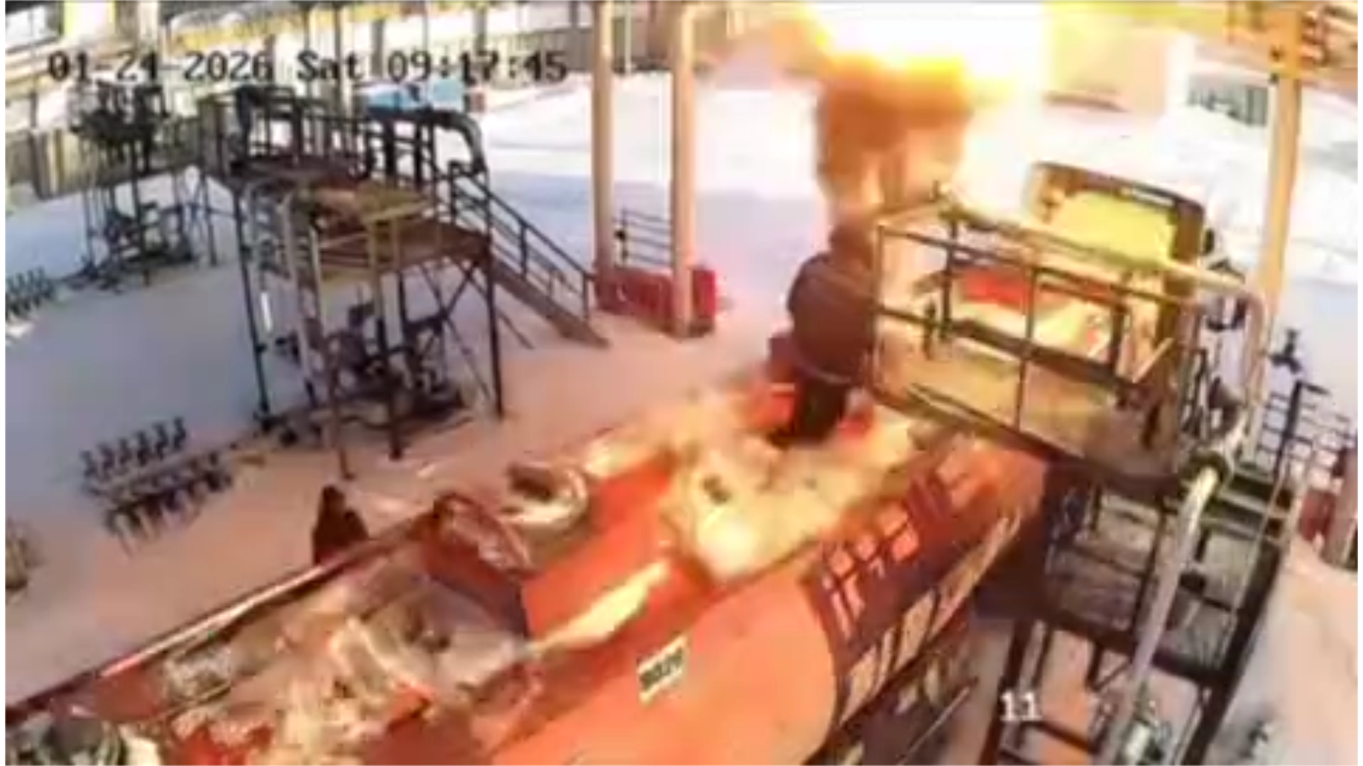 A large fuel tanker truck fully engulfed in bright orange flames at a gas station. Thick black smoke billows into the sky